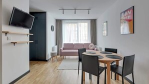 Ein modernes Wohnzimmer mit einem Flachbildfernseher, einem rosafarbenen Sofa, einem Esstisch mit Stühlen und einem großen Fenster mit Vorhängen.