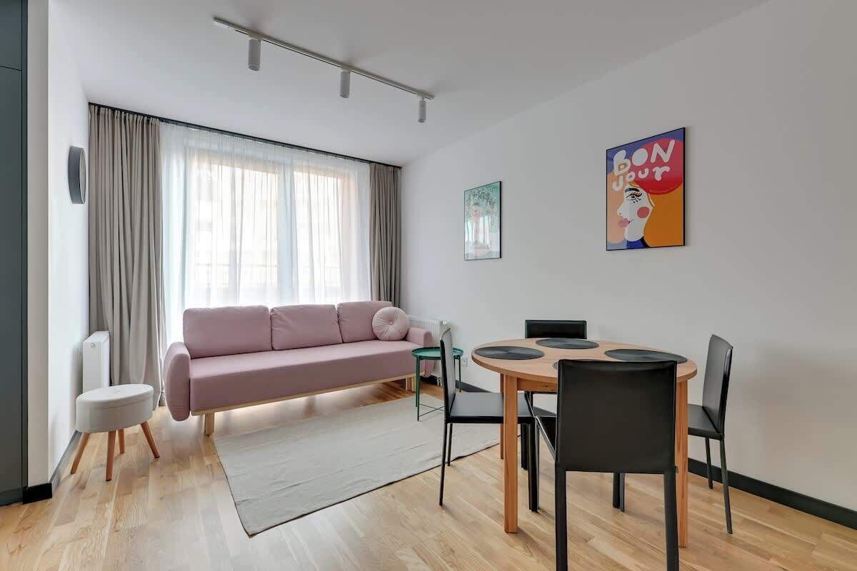 Ein modernes Wohnzimmer mit einem rosafarbenen Sofa, einem Esstisch mit Stühlen, einem Heizkörper und einem Fenster mit Vorhängen.