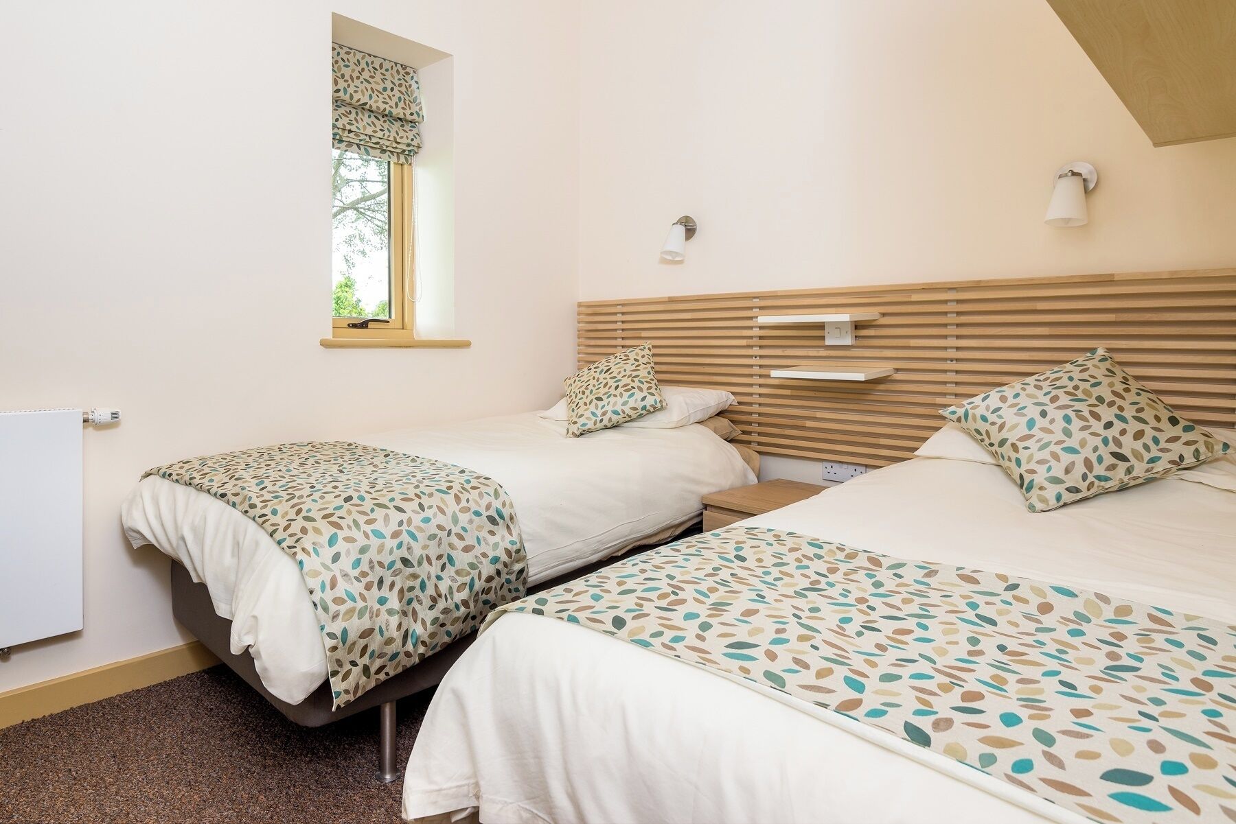 Une chambre avec deux lits, une fenêtre ornée d’une lambrequin à motifs de feuilles, un luminaire fixé au mur et une tête de lit en bois.