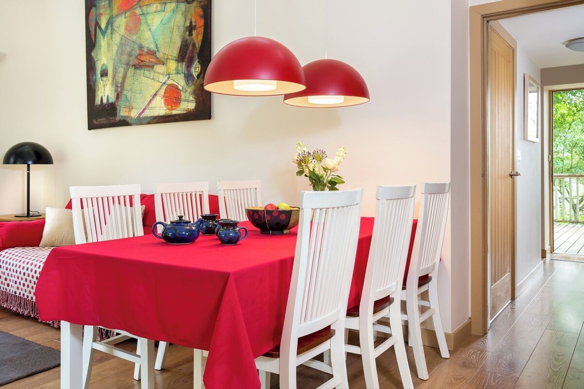 Un comedor con mantel rojo, sillas blancas, un cuadro en la pared y una puerta que da al exterior.