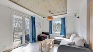 Una sala de estar moderna con un sofá, una mesa de centro y un sillón. Hay ventanas amplias con cortinas, una lámpara de techo y vistas a árboles afuera.