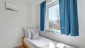 Una cama individual con ropa de cama blanca, una ventana con cortinas azules, una pequeña planta en maceta y un interruptor de luz en la pared.