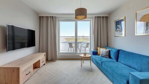 Ein modernes Wohnzimmer mit einem Flachbildfernseher, einem blauen Sofa, einem Holz-TV-Schrank, einem kleinen runden Tisch und einem großen Fenster mit Blick.