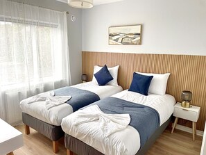 A hotel room with two beds, a bedside table, a lamp, a painting on the wall, and a window with sheer curtains. - Luxury By The Coast 6 (Galway)