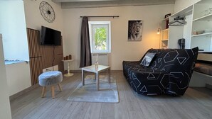 Una sala de estar con sofá, una mesita, televisor, ventana con cortinas y estante fijado a la pared.