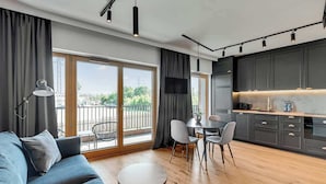 A modern living room with a sofa, dining table, and chairs, a kitchen with dark cabinets and a countertop, and large windows with a view of the city. - Nadmotławie B17A | Riverside & Gym (Gdańsk)