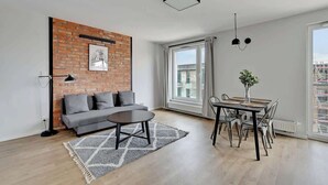 Een moderne woonkamer met een accentmuur van baksteen, een grijze bank, een ronde salontafel en een eethoek met tafel en stoelen.