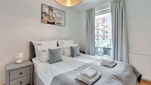 Ein modernes Hotelzimmer mit einem großen Bett, einem Nachttisch, einer Lampe, einem gerahmten Poster und Blick auf Gebäude durch das Fenster.