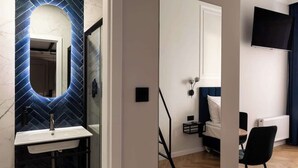 Ein modernes Hotelzimmer mit einem Badezimmer, das ein blau gefliestes Waschbecken und einen Spiegel, ein Bett mit blauer Kopfteil, einen an der Wand befestigten Flachbildfernseher und einen Holzboden zeigt.