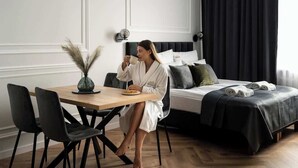 Hotellihuone, jossa on sänky, yöpöytä lampun kanssa, ruokapöytä tuoleineen sekä maljakko kuivine kasveineen.