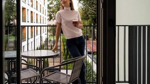 Een balkon met een tafel en stoelen, een persoon die een mok vasthoudt, en een gebouw op de achtergrond.