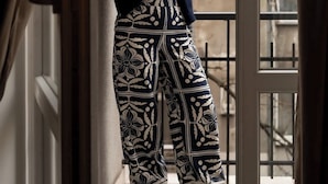 Una persona parada junto a una ventana, con pantalones y una blusa estampada, y una vista de edificios y árboles desnudos.