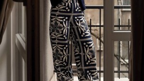 Eine Person steht an einem Fenster, draußen sind Gebäude und kahle Bäume zu sehen, die Person trägt ein Oberteil und eine Hose mit Muster.