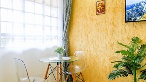 Ein Zimmer mit Glastisch, zwei Stühlen, einer Topfpflanze, einem Gemälde und einem Fenster mit Vorhängen.