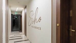 Käytävän seinässä on kyltti, jossa lukee ”Scho Residence”, ja siellä on ovi, jossa on messinkinen vetokahva, sekä kuvioitu laattalattia.