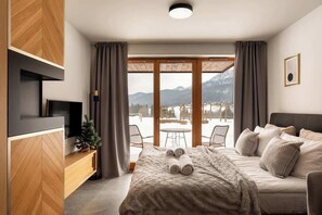 Un dormitorio con cama, televisor, aparador de madera y vistas a montañas a través de amplios ventanales.