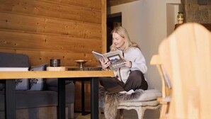 Eine Person sitzt auf einem Stuhl mit einem Fellüberwurf und liest ein Buch in einem Raum mit Holzwänden und einem Sofa.