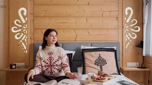 Eine Person sitzt auf einem Bett, ein Buch, ein Tablett mit Essen und ein Kissen mit Muster.