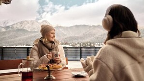 Zwei Personen sitzen an einem Holztisch mit Blick auf Berge, einer Teetasse und einem Teller Essen.
