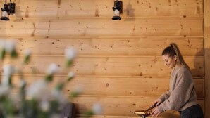 Ein Holzblockhaus-Interieur mit einer Person, die an einem Holztisch kocht, zwei an der Wand befestigten Laternen und rustikalem Ambiente.