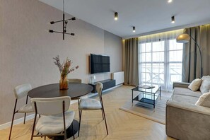 Ein modernes Wohnzimmer mit einem runden Esstisch, zwei Stühlen, einer Couch, einem an der Wand befestigten Fernseher und einem großen Fenster mit Vorhängen.