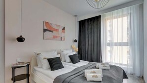 Une chambre moderne avec un lit, des tables de chevet, un luminaire suspendu, une fenêtre avec des rideaux et une œuvre d’art murale.