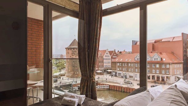 Et hotelværelse med stort vindue der kigger ud over et historisk bytorv, en seng med puder og et glasbord.