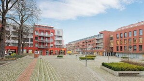Una plaza urbana moderna con edificios de ladrillo rojo, senderos pavimentados y escasa vegetación.