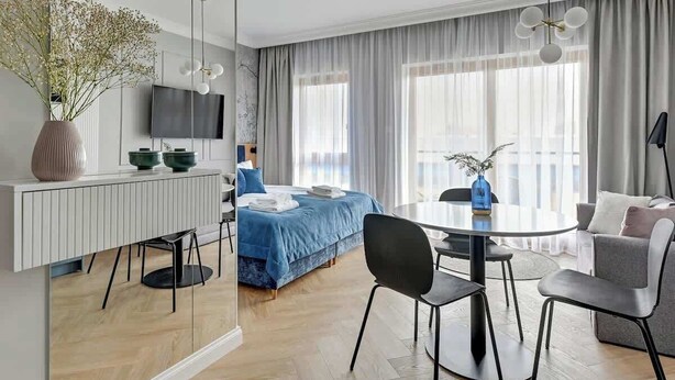 Ein modernes Hotelzimmer mit Bett, einem Esstisch mit Stühlen, einem Fernseher und einem großen Fenster mit Vorhängen.