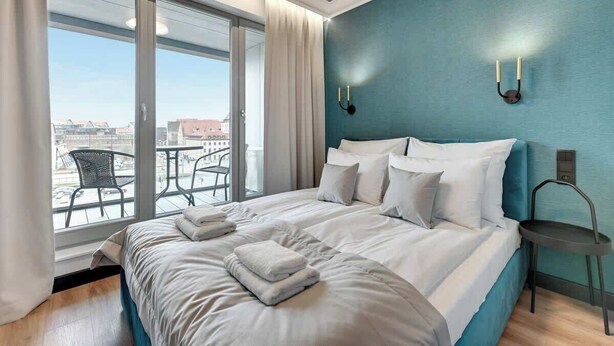 Una camera d'hotel con un letto grande, vista sulla città attraverso una porta scorrevole in vetro e un balcone con mobili da esterno.