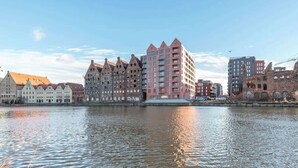 A river with buildings on the opposite bank, including a prominent pink building with a pointed roof. - Nº 75 | Pool, SPA & Gym (Gdańsk)