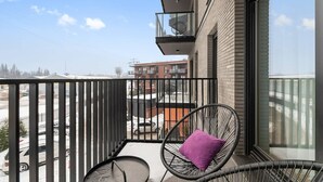 Un balcón con una silla, una mesita y una barandilla de cristal, con vistas a edificios en la distancia.