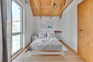Ein Schlafzimmer mit Holzboden, einem Bett mit weißer Bettwäsche, zwei gerahmten Bildern an der Wand und einem Fenster mit Vorhängen.