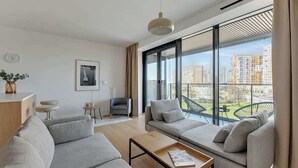A modern living room with a sofa set, a coffee table, a TV, and a large window with a city view. - Tarasy Bałtyku 68 | Seaside | Billard, Sauna & Gym (Gdańsk)