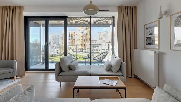A modern living room with a sofa, a coffee table, and a view of a cityscape through large windows. - Tarasy Bałtyku 68 | Seaside | Billard, Sauna & Gym (Gdańsk)