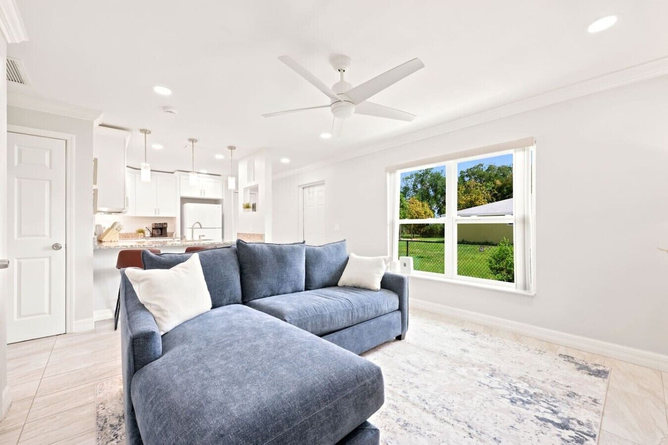 A modern living room with a gray sectional sofa, a ceiling fan, and a large window offering a view of a green landscape.