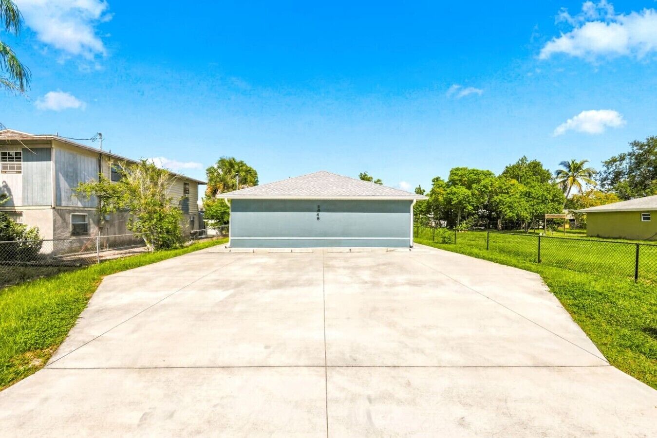 Un garage attenant à une maison, avec une allée en béton, entouré de verdure et surplombé d’un ciel bleu dégagé.