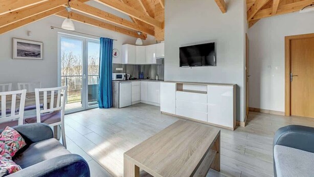 A spacious living area with a wooden ceiling, a TV mounted on the wall, a kitchen with white cabinets, and a sliding glass door leading to an outdoor area. - Dębki 6 | Rodzinny Apartament | Nad Morzem (Dębki)