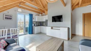 Amplia sala de estar con techo de madera, televisor montado en la pared, cocina con muebles blancos y puerta corrediza de vidrio que da a un área exterior.