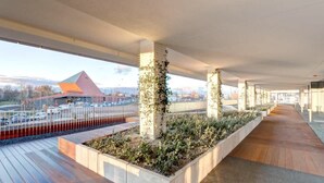 Een overdekte doorgang met betonnen pilaren, een houten terras en uitzicht op een gebouw met een rood dak.