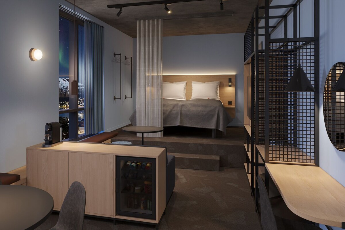 Ein modernes Hotelzimmer mit Bett, Sitzgelegenheit, kleinem Tisch und Blick auf die beleuchtete Stadt.