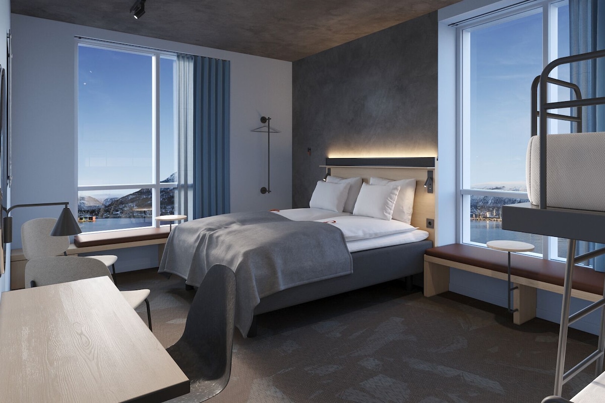Ein modernes Hotelzimmer mit Bett, Schreibtisch und Etagenbett, großen Fenstern mit Blick und einem Kopfteil mit integrierter Beleuchtung.