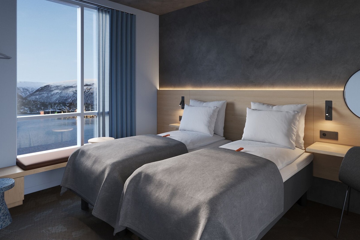 Et moderne hotelværelse med to senge, et stort vindue med udsigt til bjerge, et rundt spejl på væggen og et lille rundt bord.