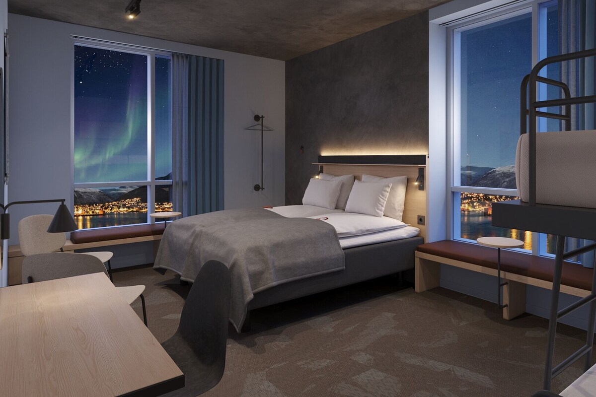 Ein modernes Hotelzimmer mit einem Bett, einem Schreibtisch, einem Sessel und einem Schlafstockbett, mit großen Fenstern und Blick auf den Nachthimmel.