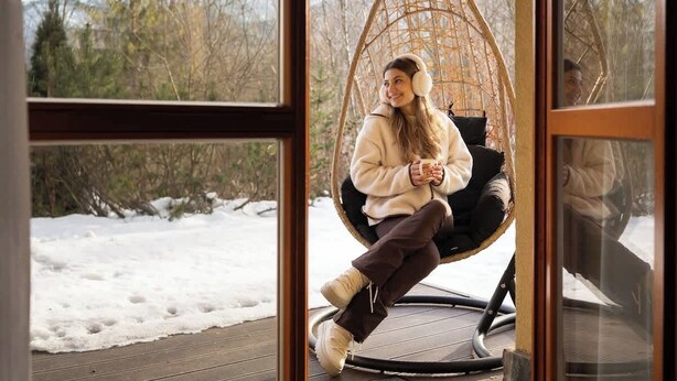 Eine Person sitzt in einem Hängestuhl vor einem Fenster mit Blick auf eine verschneite Landschaft.