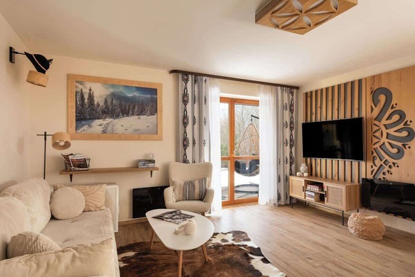 Przytulny salon w stylu górskim z wyjściem na taras. Jasna sofa, fotel, stolik kawowy i duży telewizor, a całość dopełniają naturalne drewno i regionalne akcenty. Idealne miejsce na relaks po aktywnym dniu.