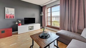 Una sala de estar moderna con un televisor de pantalla plana, un mueble blanco para televisión, una mesa de centro de madera, un sofá y un ventanal con cortinas.
