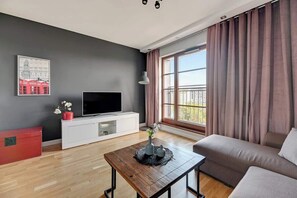Ein modernes Wohnzimmer mit Flachbildfernseher, weißem Unterhaltungsschrank, Couchtisch aus Holz, einer Couch und einem großen Fenster mit Vorhängen.