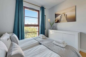 Ein Hotelzimmer mit großem Fenster, einem Bett mit weißer Bettwäsche, einem Nachttisch mit einer Vase und einem Bild an der Wand.