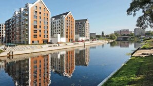 Un moderno edificio de apartamentos con su reflejo en el agua, un canal y una zona verde con un puente al fondo.